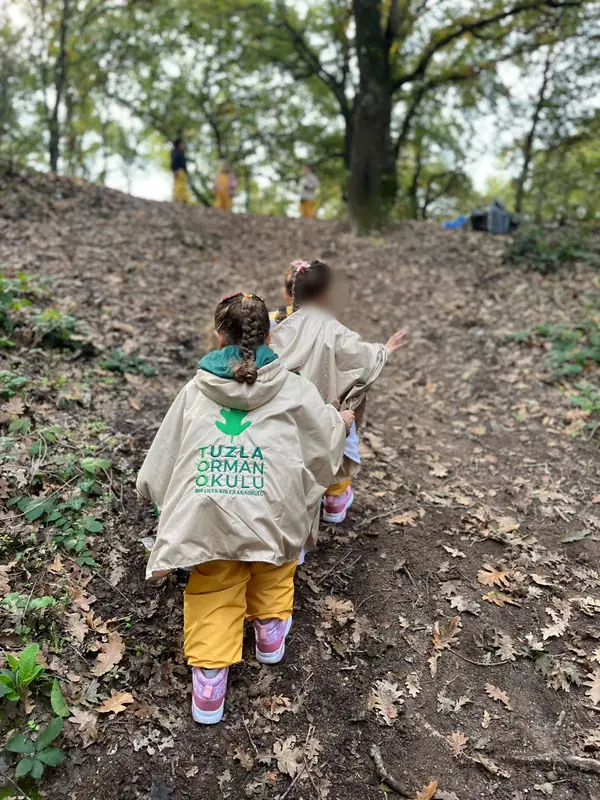 Sırtında Tuzla Orman Okulu logolu bej panço olan iki çocuk eğimli orman yolunda yürüyor, arkadan